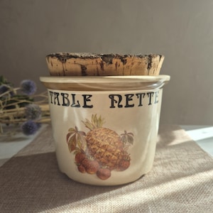 Puede incluir: Un recipiente de cerámica de color crema con una tapa de corcho gruesa y texturizada. El frente presenta el texto negro "TABLE NETTE" sobre una ilustración colorida de una piña rodeada de fresas y hojas verdes sobre un fondo beige claro.