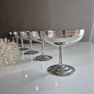 Peut inclure: Ensemble de cinq coupes à champagne argentées. Les verres ont un large bol peu profond, une tige fine et une base ronde. Les verres sont disposés en rangée, reflétant la lumière. L'arrière-plan est gris neutre.