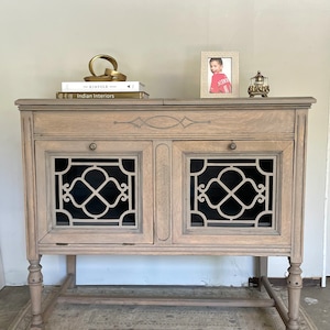 May include: A light brown wooden cabinet with two doors featuring black geometric designs. The cabinet has a decorative top, two drawers, and is adorned with a gold sculpture, books, a framed photo, and a small decorative object.