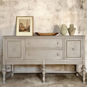 Peut inclure: Buffet peint en gris avec deux tiroirs et deux portes d'armoires. Le buffet a des pieds tournés et une garniture décorative. Sur le dessus se trouvent un tableau encadré, un bol en bois et deux vases en céramique. Le fond est un mur texturé.