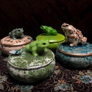 May include: A selection of ceramic trinket boxes, each topped with a frog ornament. The boxes display diverse colours and textures, including speckled and mottled finishes. One box is decorated with a butterfly.