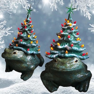 Può includere: Due statuine di alberi di Natale in ceramica sopra sculture di rane. Gli alberi sono verdi con luci rosse e gialle e una stella verde. Le rane sono di colore verde e blu screziato, con una finitura strutturata. Lo sfondo è una scena invernale innevata.