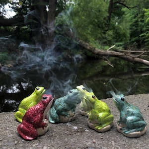 Può includere: Un gruppo di sei statuette di rana in ceramica nei toni del verde, rosso e verde acqua, disposte su una superficie di pietra. Volute di fumo si alzano dalle rane, creando un effetto etereo. Lo sfondo presenta un ambiente naturale sfocato con acqua e alberi.