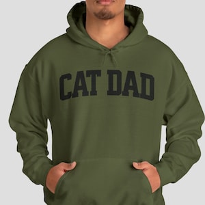 Military Green colored pullover hoodie with Cat Dad printed in an arch on the front in black.  Varsity athletic block style font printed on the hooded sweatshirt.