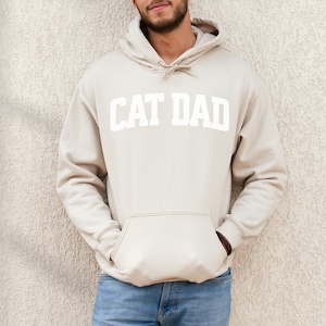 Sand colored pullover hoodie with Cat Dad printed in an arch on the front in white.  Varsity athletic block style font printed on the hooded sweatshirt.