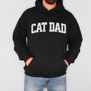 Black colored pullover hoodie with Cat Dad printed in an arch on the front in white.  Varsity athletic block style font printed on the hooded sweatshirt.