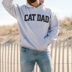 Sport Gray colored pullover hoodie with Cat Dad printed in an arch on the front in black.  Varsity athletic block style font printed on the hooded sweatshirt.