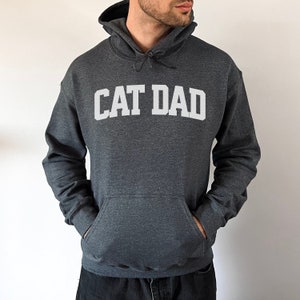 Dark heather colored pullover hoodie with Cat Dad printed in an arch on the front in white.  Varsity athletic block style font printed on the hooded sweatshirt.