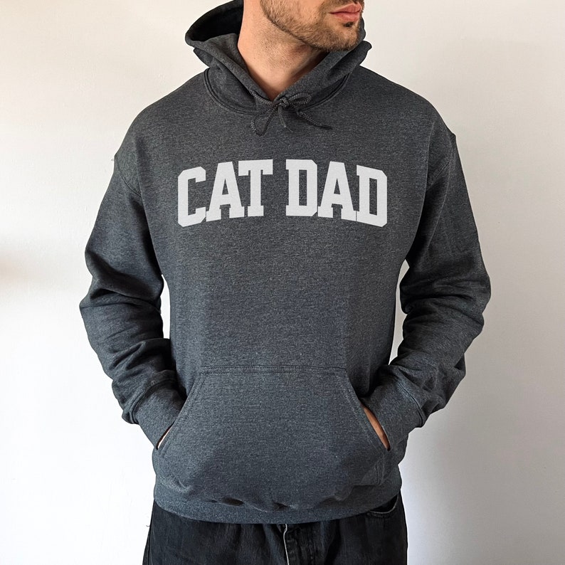 Dark heather colored pullover hoodie with Cat Dad printed in an arch on the front in white.  Varsity athletic block style font printed on the hooded sweatshirt.