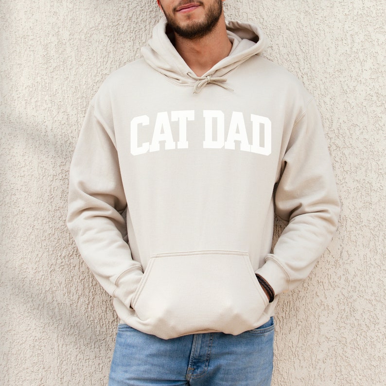 Sand colored pullover hoodie with Cat Dad printed in an arch on the front in white.  Varsity athletic block style font printed on the hooded sweatshirt.