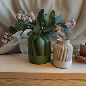 Op de afbeelding: Twee keramische vazen met een geribbeld ontwerp. De vaas links is groen en de vaas rechts is beige. Beide vazen zijn gevuld met roze bloemen.