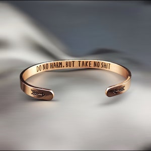 May include: Rose gold cuff bracelet with the engraved text "DO NO HARM, BUT TAKE NO SHIT". The bracelet features an arrow design on each end. The open design allows for easy wear.