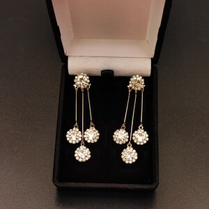 May include: A pair of gold dangle earrings with three clear crystal stones on each earring. The earrings are in a black velvet box.