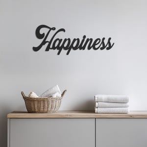 Może przedstawiać: Czarna metalowa dekoracja ścienna z napisem "Happiness" w ozdobnym, pochylonym piśmie. Dekoracja wisi na białej ścianie nad drewnianą półką z wiklinowym koszem i białymi ręcznikami.