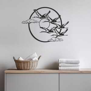 May include: Black metal wall art featuring a circle with a sun and a flock of birds flying in formation. The art is mounted on a white wall above a wooden shelf with a wicker basket and towels.