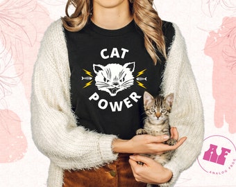 Puede incluir: Una camiseta negra con un gráfico de gato blanco y el texto "CAT POWER" en letras blancas. El gato tiene rayos amarillos que salen de sus orejas y una espina de pescado en la boca.