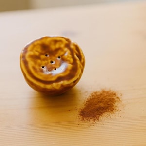 Pode incluir: Um polvilhador de cerâmica redondo com um esmalte brilhante em tons de castanho e uma série de pequenos orifícios na parte superior. Ao lado, encontra-se um pequeno monte de pó fino castanho-avermelhado, semelhante a canela moída.