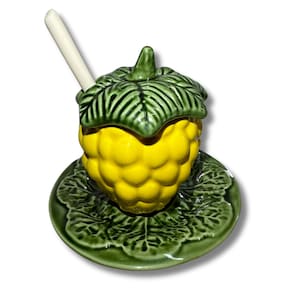Pode incluir: Um pote de mel de cerâmica amarelo em forma de cacho de bagas com uma tampa de folha verde e uma pequena colher branca. O pote está sobre um pires de cerâmica verde com um desenho de folhas.