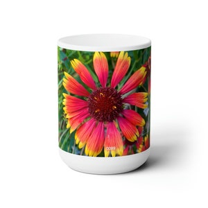 May include: A white ceramic mug with a colorful flower design. The flower has red, orange, and yellow petals with a dark red center. The background is green.
