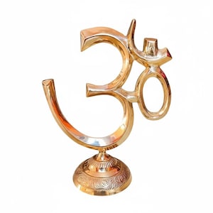 Könnte beinhalten: Eine goldfarbene Om-Symbol-Skulptur. Die Skulptur besteht aus Metall und hat einen detaillierten, verzierten Sockel. Das Om-Symbol ist ein heiliger Klang und eine spirituelle Ikone im Hinduismus, Buddhismus und Jainismus. Die Skulptur ist ein Dekorationsartikel.