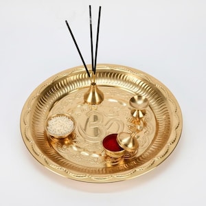 May include: A gold-coloured puja thali with detailed embossed patterns. The thali contains a small bowl of rice, a container of red powder, a small bell, and a holder with three incense sticks. A traditional item for religious ceremonies.