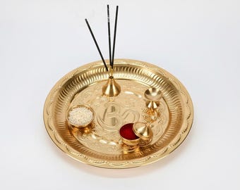May include: A gold-colored puja thali with intricate embossed designs. The thali holds a small bowl of rice, a container of red powder, a small bell, and a holder with three incense sticks. A traditional item for religious ceremonies.