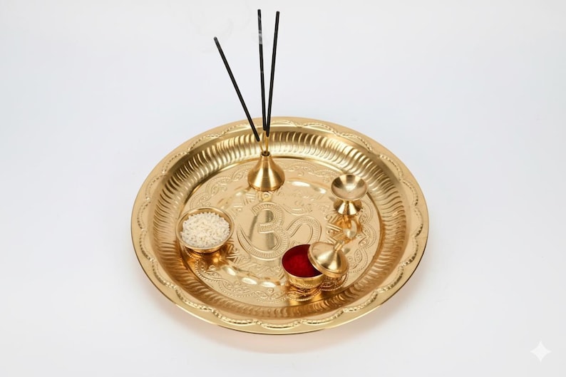 May include: A gold-coloured puja thali with detailed embossed patterns. The thali contains a small bowl of rice, a container of red powder, a small bell, and a holder with three incense sticks. A traditional item for religious ceremonies.