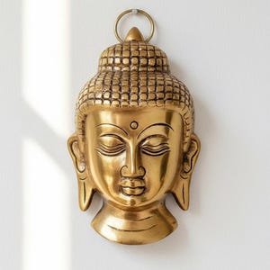 Può includere: Una placca da parete dorata a forma di testa di Buddha con ushnisha testurizzato, occhi chiusi e un piccolo bindi circolare sulla fronte. La scultura presenta lobi delle orecchie allungati e un anello di metallo superiore per il fissaggio.
