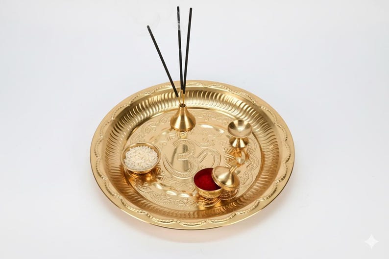 May include: A gold-coloured puja thali with detailed embossed patterns. The thali contains a small bowl of rice, a container of red powder, a small bell, and a holder with three incense sticks. A traditional item for religious ceremonies.