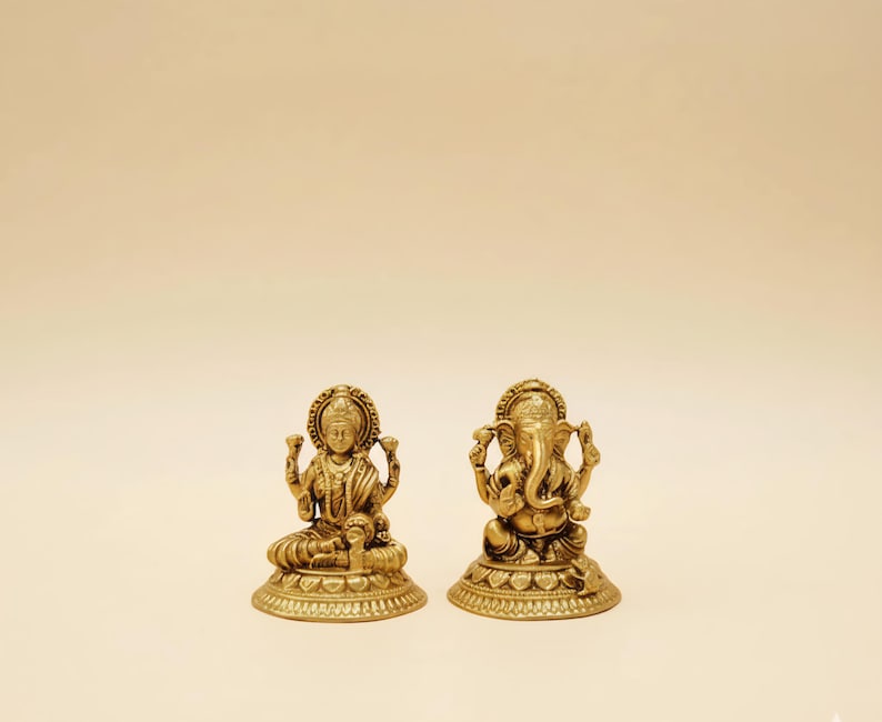 Pode incluir: Duas est&aacute;tuas douradas de divindades hindus. Uma est&aacute;tua retrata Lakshmi, a deusa da riqueza, sentada com v&aacute;rios bra&ccedil;os. A outra est&aacute;tua &eacute; de Ganesha, o deus com cabe&ccedil;a de elefante, tamb&eacute;m sentado. Ambas as est&aacute;tuas est&atilde;o em um fundo bege claro.