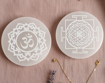 Puede incluir: Dos posavasos de cristal de selenita blancos con diseños grabados, uno con un símbolo Om y el otro con un patrón geométrico. Cristales de cuarzo rosa y un collar de oro con un colgante de cristal transparente también están presentes. Los artículos están dispuestos sobre una superficie rosa.