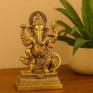 Op de afbeelding: Een gouden messing beeld van de hindoe&iuml;stische godheid Ganesha, zittend op een rat. Het beeld heeft ingewikkelde details, waaronder Ganesha's olifantenkop, meerdere armen en sierlijke sieraden. De basis is rechthoekig en de achtergrond bevat een plant in een witte pot.