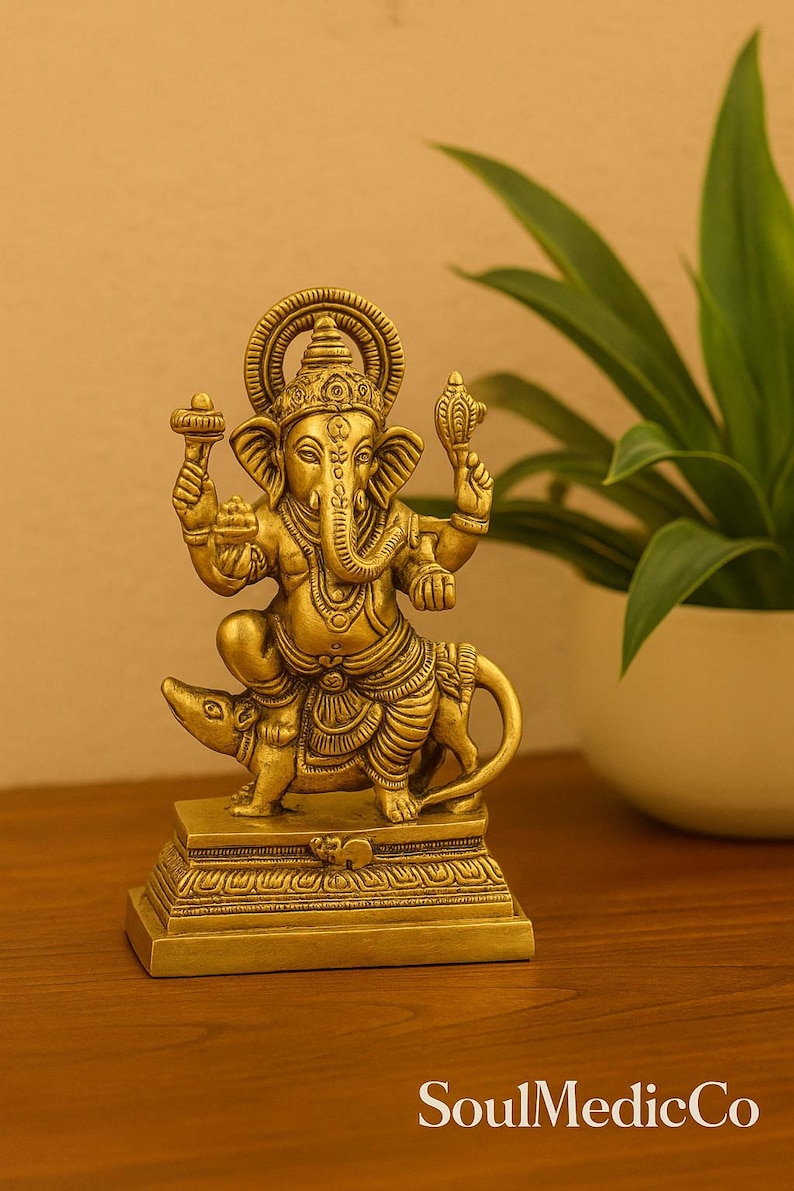 Op de afbeelding: Een gouden messing beeld van de hindoe&iuml;stische godheid Ganesha, zittend op een rat. Het beeld heeft ingewikkelde details, waaronder Ganesha's olifantenkop, meerdere armen en sierlijke sieraden. De basis is rechthoekig en de achtergrond bevat een plant in een witte pot.