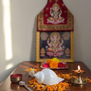 May include: A traditional Hindu altar setup featuring a framed deity image, a white conch shell, a small brass lamp with a lit flame, and a small bowl of red powder. The scene is adorned with orange and yellow flower petals.