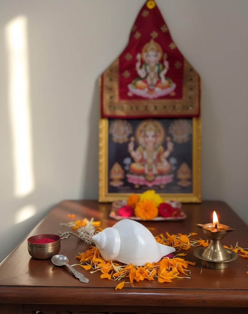 May include: A traditional Hindu altar setup featuring a framed deity image, a white conch shell, a small brass lamp with a lit flame, and a small bowl of red powder. The scene is adorned with orange and yellow flower petals.