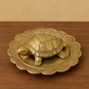 Op de afbeelding: Een messing schildpadfiguur rust op een decoratieve messing plaat met een geschulpte rand. De schildpad heeft een gedetailleerd schild en bevindt zich in het midden van de plaat. De plaat en schildpad zijn goudkleurig.
