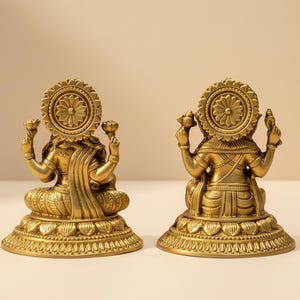Pode incluir: Duas est&aacute;tuas douradas de uma divindade, vistas por tr&aacute;s. Cada est&aacute;tua tem uma aur&eacute;ola circular, roupas ornamentadas e est&aacute; sentada em uma base detalhada. As est&aacute;tuas est&atilde;o em uma superf&iacute;cie clara contra um fundo neutro.