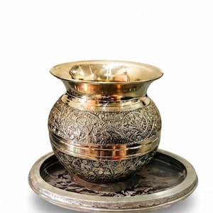 May include: A decorative metal pot with intricate floral patterns, sitting on a matching round tray. The pot has a wide, flared rim and a bulbous body. The metal has a shiny, polished finish, and the tray has a detailed edge.