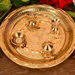 May include: A gold-coloured metal puja plate with intricate embossed designs and small decorative bowls. The plate features an Om symbol in the centre and is surrounded by floral accents.