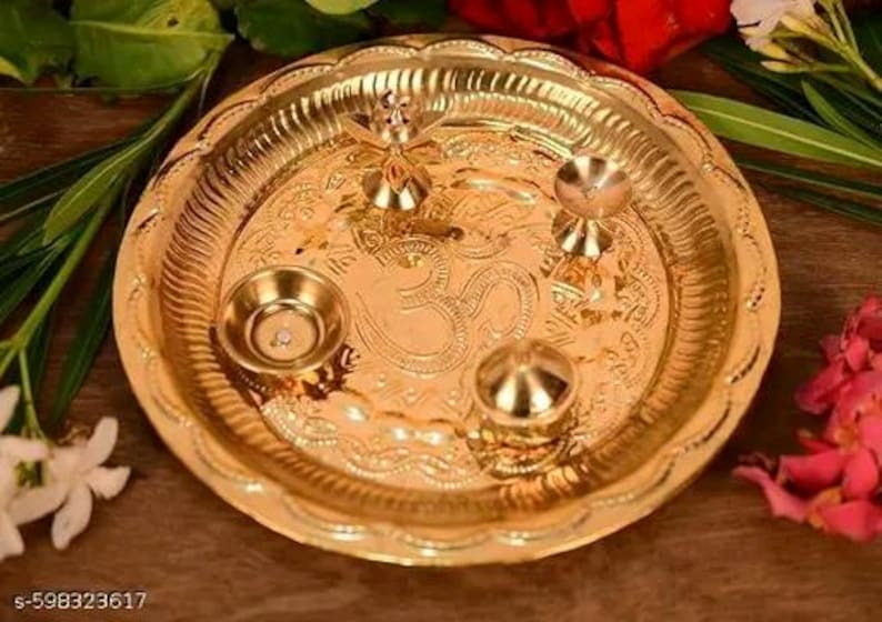 May include: A gold-coloured metal puja plate with intricate embossed designs and small decorative bowls. The plate features an Om symbol in the centre and is surrounded by floral accents.