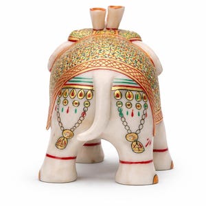 Puede incluir: Una figura decorativa de elefante blanco con intrincados detalles pintados en oro, rojo y verde. El elefante est&aacute; adornado con una manta estampada y cadenas decorativas. La figura tiene una trompa y colmillos detallados.
