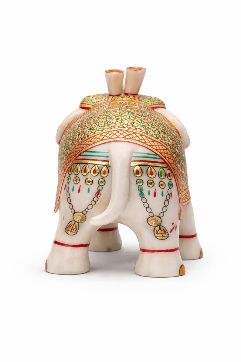 Puede incluir: Una figura decorativa de elefante blanco con intrincados detalles pintados en oro, rojo y verde. El elefante est&aacute; adornado con una manta estampada y cadenas decorativas. La figura tiene una trompa y colmillos detallados.
