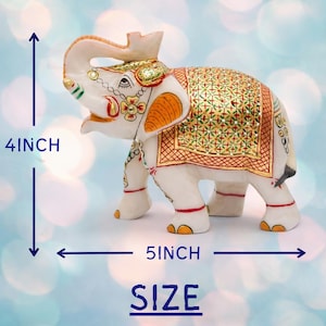 Puede incluir: Figura de elefante blanco, 10 cm de alto y 13 cm de largo, con detalles pintados en oro y naranja. El elefante tiene una manta decorativa y est&aacute; sobre un fondo azul borroso. La palabra "SIZE" es visible.
