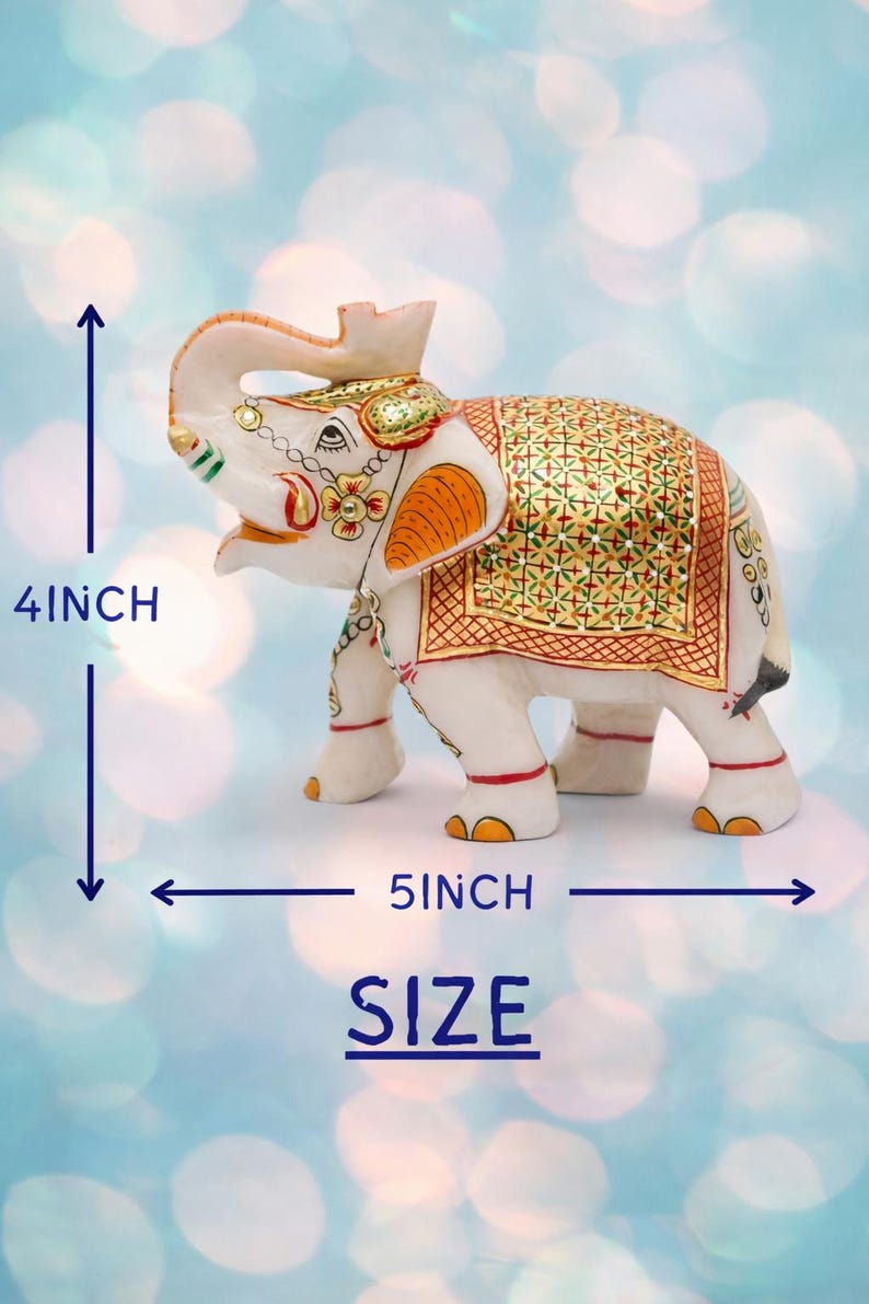 Puede incluir: Figura de elefante blanco, 10 cm de alto y 13 cm de largo, con detalles pintados en oro y naranja. El elefante tiene una manta decorativa y est&aacute; sobre un fondo azul borroso. La palabra "SIZE" es visible.