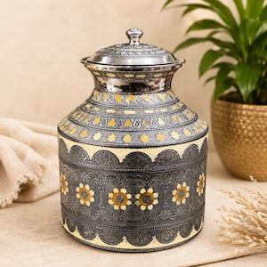 May include: A cylindrical metal vessel with a lid, featuring a silver and cream finish with intricate embossed floral patterns and yellow accents. The design includes a repeating scalloped border and detailed engravings across the entire surface.