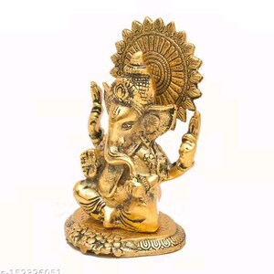 May include: A metallic gold-toned figurine of the deity Ganesha seated in a cross-legged position. The figure features an elephant head, multiple arms, and an ornate circular halo behind the head, all set upon a textured, circular base.