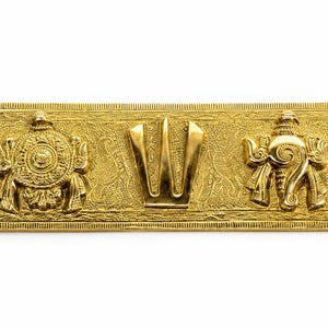 May include: A rectangular brass plaque featuring a central U-shaped symbol flanked by a circular wheel emblem on the left and a conch shell emblem on the right. The metal surface has a textured background and four small holes at the corners.