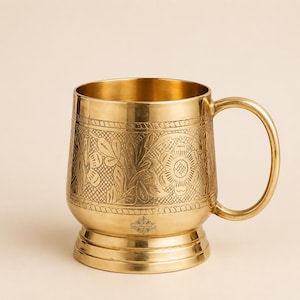 Könnte beinhalten: Ein Becher aus poliertem Messing mit einem komplizierten, eingravierten Blumenmuster in der Mitte, einem glatten Henkel und einem breiten, gestuften Boden. Der Text SoulMedicCo ist in kleinen Buchstaben unten am Blumenmuster eingraviert.