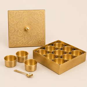 May include: A square gold-toned metal box with a textured lid featuring a circular pattern and a small knob. Inside the box are twelve small, round, gold-toned cups. Three additional cups and a small gold-toned spoon are placed outside the box.