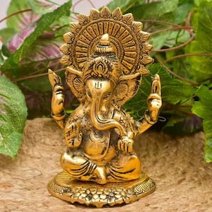 May include: A metallic gold-toned statue of the deity Ganesha, depicted in a seated position with an elephant head, large ears, and a trunk curved to the left. The figure features an ornate circular halo behind the head and detailed engravings.